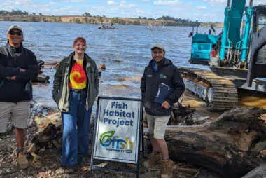 Three people standing on the riverbank with a Fish habitat restoration sign and then there are logs and a machine in the back