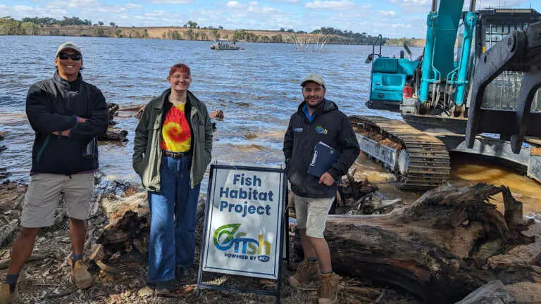 Three people standing on the riverbank with a Fish habitat restoration sign and then there are logs and a machine in the back