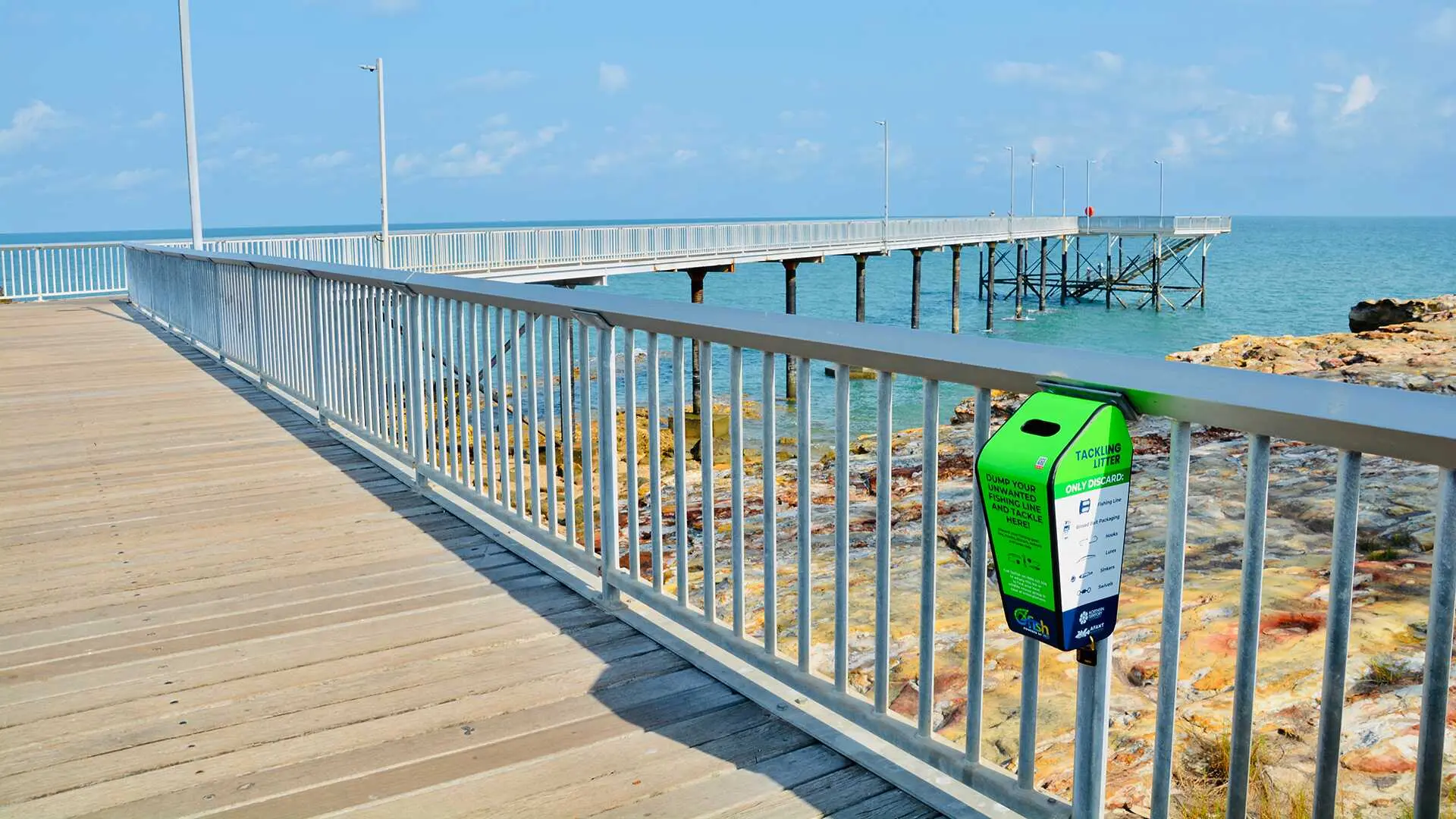 A green bin attached to a rail along a jetty with a blue sky and water in the background