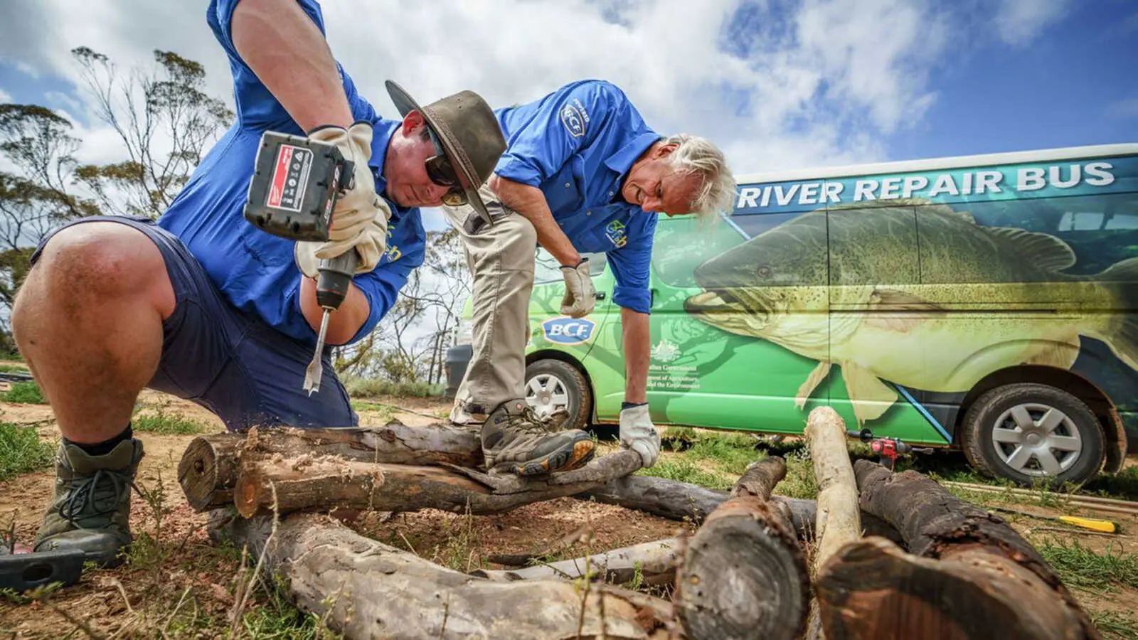 Two men crouched over some logs with a drill and a River Repair Bus in the background