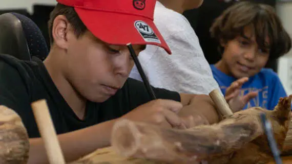 A close up of an Indigenous boy wearing a red cap drawing on a log with a pencil