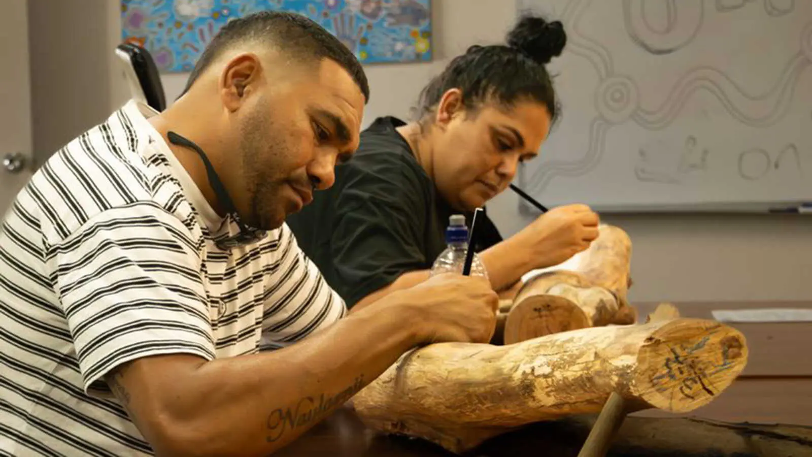 An Indigenous woman and man sitting at a table drawing on logs with pencils