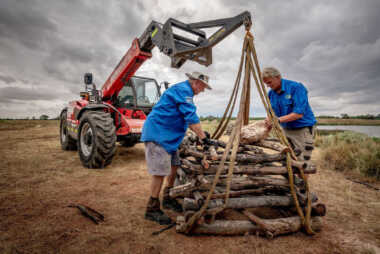 two men securing logs to be transferred by a machine in the background