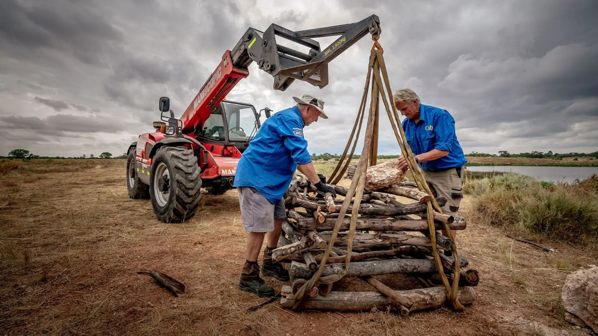 two men securing logs to be transferred by a machine in the background