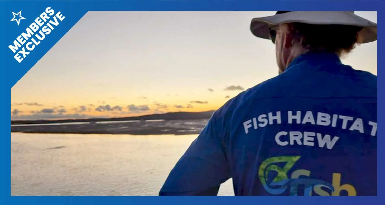 The back of a man in a blue shirt with Fish Habitat Crew written on the back, looking out over the sunset on the water