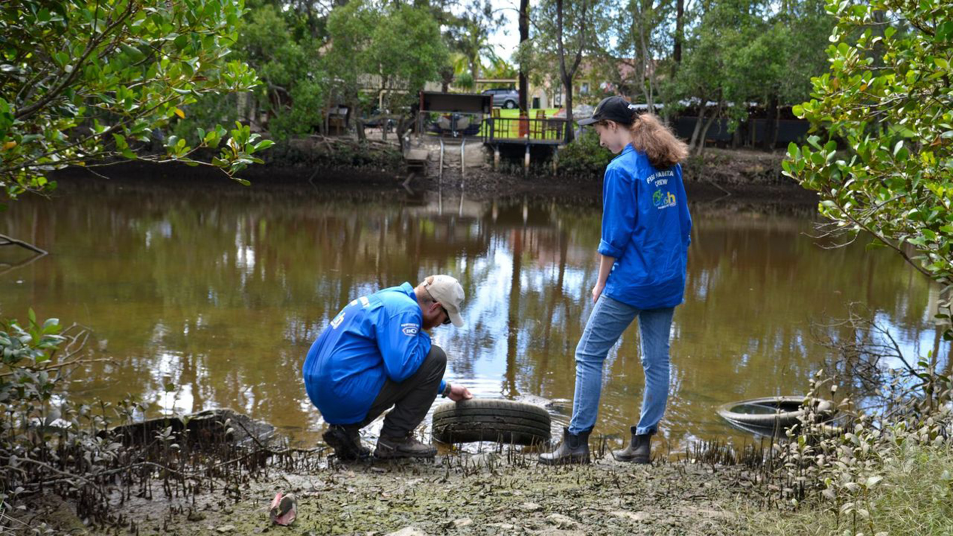 two people in blue shirts in front of a creek, one bending down pulling a tyre out of the water and the other person standing looking over them